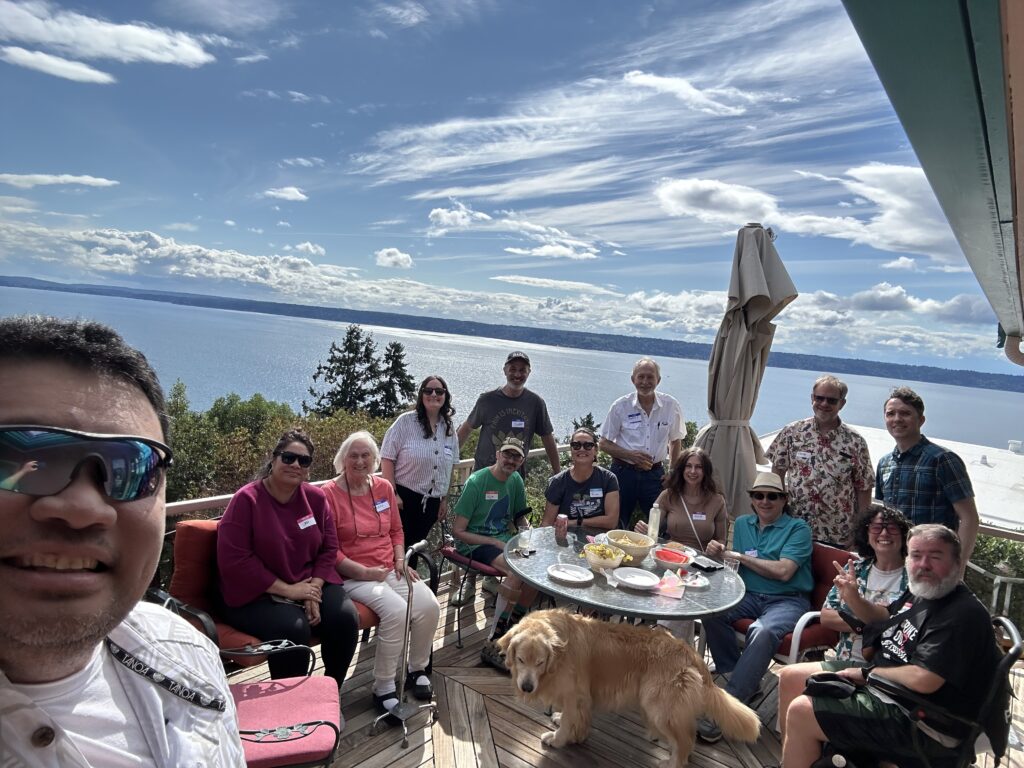 About a dozen people seated around an outdoor table with a beautiful view of water and blue sky with wispy clouds behind them.